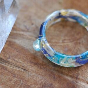 Rainbow moonstone rounded stone set in a clear resin band made of blue and white cornflowers, 24k gold flakes and blue skeleton leaves.