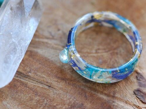 Rainbow moonstone rounded stone set in a clear resin band made of blue and white cornflowers, 24k gold flakes and blue skeleton leaves.